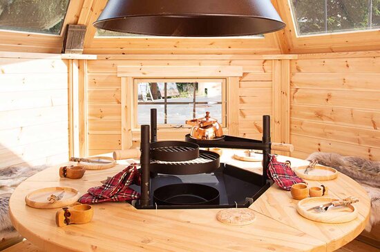 Sonnendurchfluteter Innenraum einer Grillkota von Finnhaus Wolff mit Grill in der Mitte und Essensgedeck aus Holz.