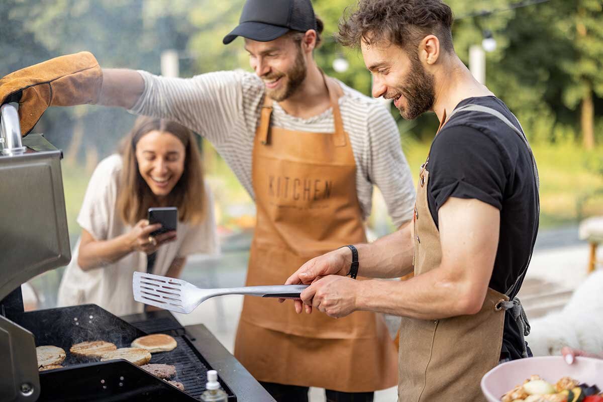 2 Männer grillen Burger, eine Frau filmt das Szenario mit Smartphone.